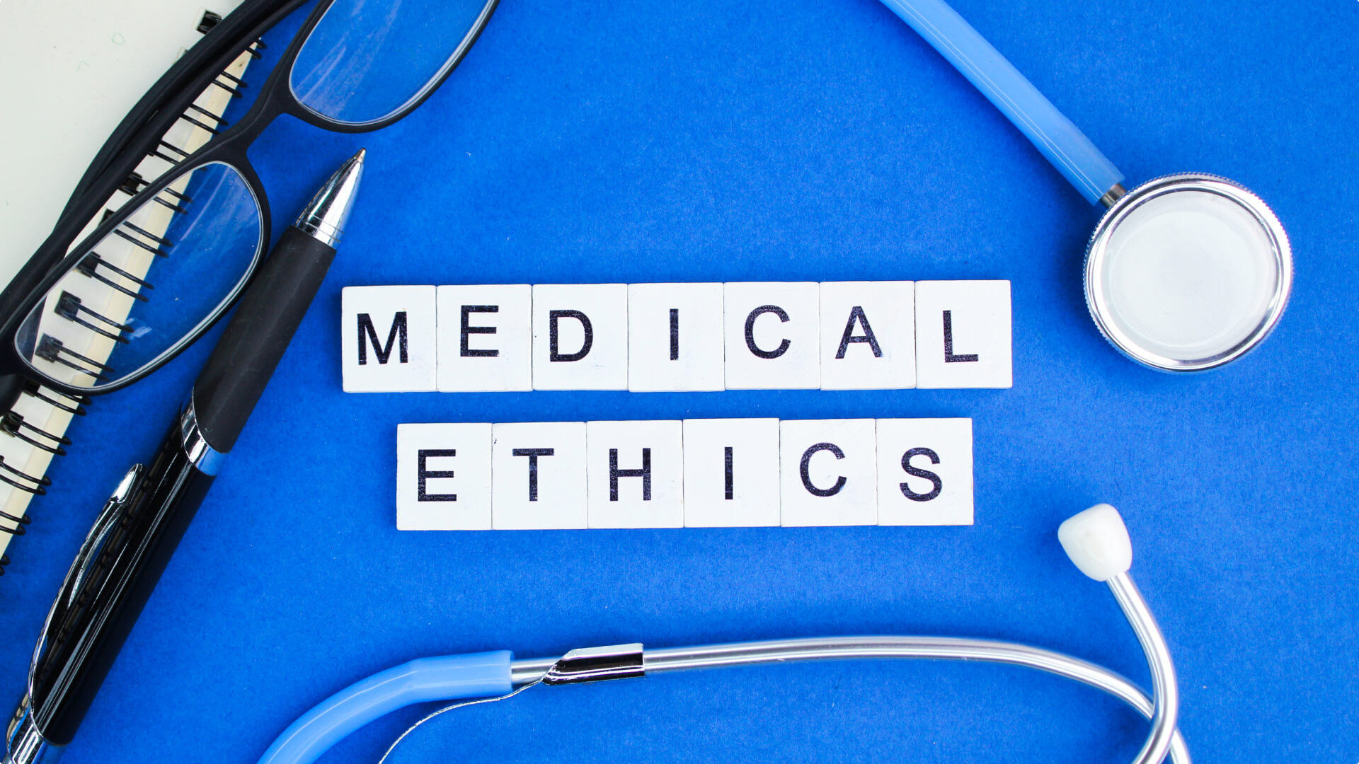 Stethoscope, eyeglasses, pen, and the words 'MEDICAL ETHICS' spelled out on tiles on a blue background, representing the ethical considerations in healthcare.